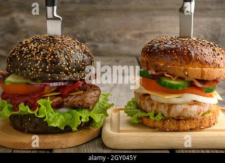 Zwei Cheeseburger mit Gemüse und Fleischsteak auf einem Holzbrett, ein Sandwich wird mit einem Messer durchbohrt Stockfoto