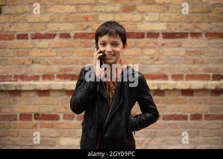 Hoto des Teenagers im Alter von 11-12 Jahren spricht auf dem Smartphone vor dem Hintergrund einer Backsteinmauer in der Stadt, lächelt auf seinem Gesicht Stockfoto