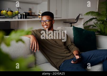 Junger Erwachsener schwarzer Mann, der sich auf dem Sofa mit dem Mobiltelefon in einer modernen Wohnung entspannt Stockfoto