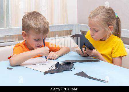 Junge und Mädchen schneiden zu Hause Halloween Fledermäuse mit einer Schere aus schwarzem Papier. Kinder machen Fledermäuse aus schwarzem Papier. Heimwerken zu Hause. Startseite Halloween Urlaub c Stockfoto