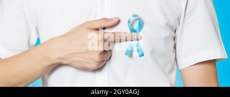 Men's Healthcare Konzept - Nahaufnahme der männlichen Hand zeigt auf hellblaues Band für Prostatakrebs auf der linken Brust auf blauem Hintergrund. Bannergröße mit Stockfoto