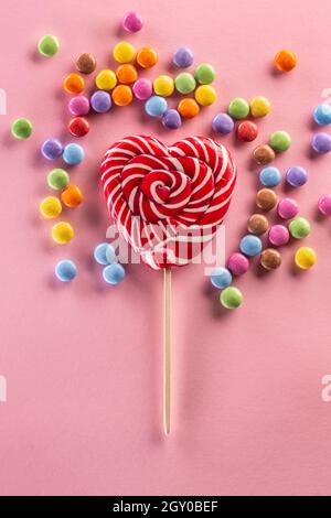 Herzähnlich gestreifter roter Lollipop, umgeben von süßen, farbenfrohen Bonbons, die auf dem rosa Hintergrund liegen. Stockfoto