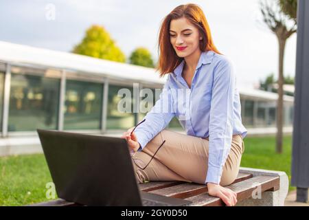 Junge rothaarige kaukasische Geschäftsfrau im Park lächelt in den Laptop Stockfoto
