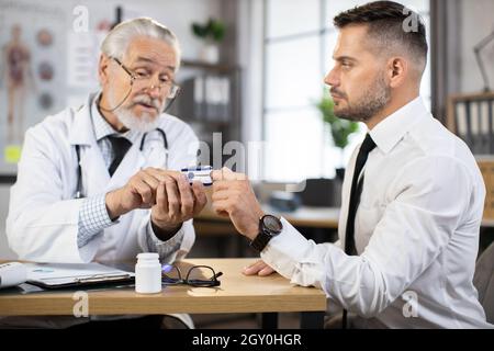 Kompetenter leitender Arzt, der das Fingerpulsoximeter zum Testen des Sauerstoffgehalts im Blut von kaukasischen Männern in Geschäftskleidung verwendet. Konzept von Menschen, Untersuchung und Behandlung. Stockfoto