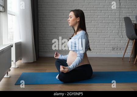 Schöne Schwangerin, die zu Hause, horizontal trainiert. Sitzt in der Lotusposition und meditiert. Stockfoto