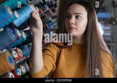 Eine junge Näherin in einer Werkstatt steht in der Nähe eines Standes mit Fäden verschiedener Farben. Die Wahl des Nahtmaterials zum Nähen eines Hemdes. Stockfoto