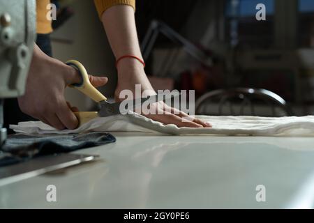 Damenhänden schneiden Stoff auf einem Muster mit Schneiderschere auf einem weißen Tisch. Ein Schneidermeister bei der Arbeit in einem Nähstudio. Stockfoto