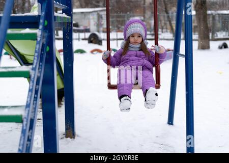 Kinderspielplatz im Winter. Ein kleines Mädchen fährt während eines Schneefalls auf einer Schaukel. Stockfoto