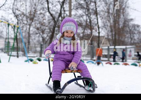 Porträt eines glücklichen Mädchens. Ein Kind im Winter ist in einer violetten hellen Jacke gekleidet, um während der Sommerferien Schlittenfahren zu gehen. Stockfoto