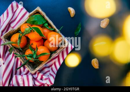 Draufsicht frisch reife Mandarinen, Clementine, Mandarine mit grünen Blättern in der Strohschale auf gestreifter Serviette und dunkelblauem Steinhintergrund mit hellem Stockfoto