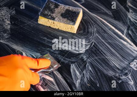 Der orangefarbene Handschuh verwendet einen Metallschaber, um das Kochfeld mit dem Reinigungsmittel zu reinigen. Selektiver Fokus. Blick von oben. Ein gelber Schwamm ruht Stockfoto