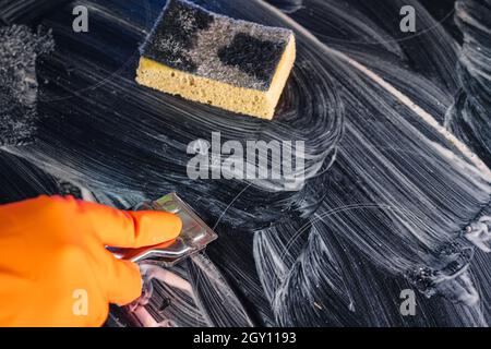 Der orangefarbene Handschuh verwendet einen Metallschaber, um das Kochfeld mit dem Reinigungsmittel zu reinigen. Selektiver Fokus. Blick von oben. Ein gelber Schwamm ruht Stockfoto