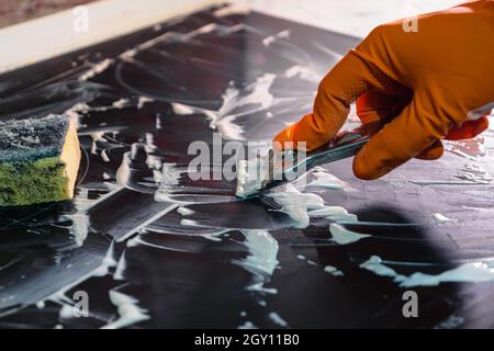 Eine Hand in einem orangefarbenen Handschuh reinigt das Kochfeld mit einem Metallschaber. Selektiver Fokus. Das Konzept der Hausreinigung eines Elektroofens Stockfoto
