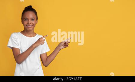 Frohes lächelndes, schwarzes Mädchen im weißen T-Shirt, das die Finger zur Seite zeigt, um den freien Raum zu schaffen Stockfoto