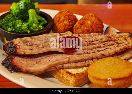 Scheiben Brisket mit Brot, Maismuffin, Hush Welpen, und Seite von Brokkoli auf einem weißen Teller Stockfoto