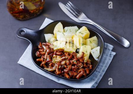 Pfifferlinge mit gekochten Kartoffeln, Zwiebeln, Dill und Öl in einer kleinen schwarzen Pfanne. Traditionelles russisches Gericht. Schwarzer Hintergrund, Nahaufnahme, Kamera-Zo Stockfoto