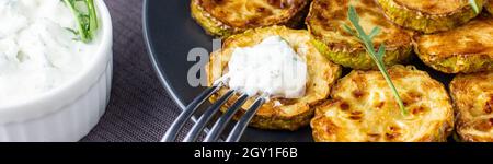 Banner von gebratenen Zucchini-Scheiben mit saurer Rahmsoße auf dem Teller. Gebratene Zucchini mit Kraut zum Abendessen auf natürlichem braunen Holzhintergrund. Top-wlan Stockfoto