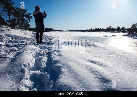 Frau in warmer Kleidung, die mit ihrem Phono die wunderschöne Winterlandschaft des schwedischen Archipels fotografiert, während sie im Schnee steht. Stockfoto