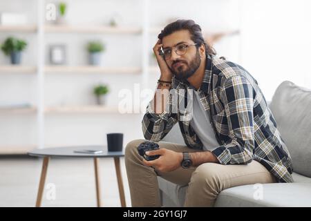 Gelangweilter indischer Kerl, der mit Joystick auf der Couch sitzt Stockfoto
