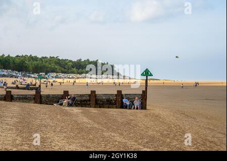 Menschen, die das Strandleben genießen, Paare, die neben einer hölzernen Groyne mit Strandhütten und Wäldern als Hintergrund sitzen, Wells Next the Sea, Norfolk, England. Stockfoto