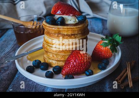 Nahaufnahme des Stapels von Pfannkuchen mit Honig, Erdbeere, Blaubeere und Peitschencreme auf dem Teller, leckeres Dessert mit Milch zum Frühstück. Stockfoto