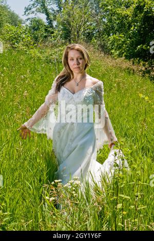 Schöne Frau mit langen braunen Haaren in einem fließenden weißen Kleid auf einer Wiese unter hohen Gräsern Stockfoto