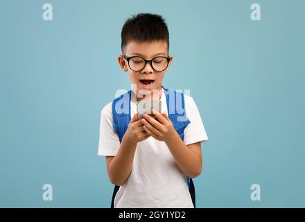 Aufgeregt Asiatische Schuljunge Mit Rucksack Mit Smartphone Über Blauen Hintergrund Stockfoto