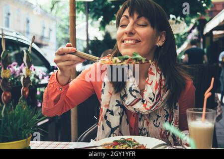 Lächelnde junge Frau, die gerne italienische vegetarische Pizza und Kaffee Latte zum Mittagessen im italienischen Restaurant im Freien isst Stockfoto