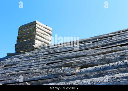 Eine Nahaufnahme eines grauen und verwitterten, gespaltenen Holzdachs und Schornsteins. Blauer Himmel im Hintergrund. Stockfoto
