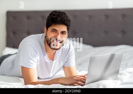 Immer online. Junger arabischer Mann, der entfernt am Laptop arbeitet, auf dem Bett liegt und vor der Kamera lächelt, freier Platz Stockfoto