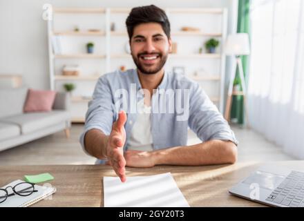 Beschäftigungskonzept. Arabischer Geschäftsmann streckt die Hand zum Händeschütteln, begrüßt die Kamera, sitzt am Arbeitsplatz zu Hause Stockfoto