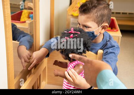 Bildung Preschool Junge im Alter von 3-4 Jahren im Familienbereich hält Puppe zum Spiegel und zeigt auf Reflexion, während die Hand des Lehrers von der Seite nach innen zeigt und das fa trägt Stockfoto