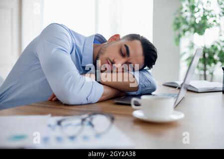 Müde arabischer Geschäftsmann, der vor dem Laptop auf seinem Schreibtisch schläft, überfordert ist und sich im modernen Büro erschöpft fühlt Stockfoto