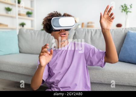 Afroamerikanischer Teenager-Typ im Virtual-Reality-Headset, der imaginären Bildschirm berührt und den Cyberspace zu Hause erkundet Stockfoto