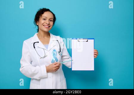 Wunderschöne Ärztin in weißem medizinischen Kleid und blauem Bewusstseinsband lächelt toothy Lächeln Blick auf die Kamera und zeigt eine Kopie Platz auf leeren leeren Pap Stockfoto