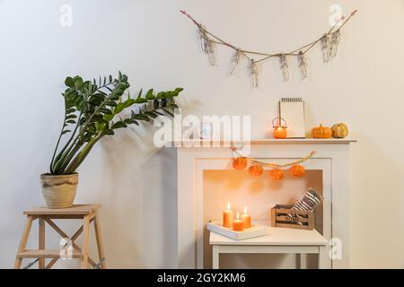 Rahmen mit Schild 'Happy Halloween'. Zamioculcas Pflanzen im Tontopf auf dem Stuhl. Girlande von Skeletten hängt an der Wand. Girlande aus Kunststoff-Kürbissen hängt von Stockfoto