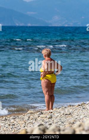 Eine übergewichtige Dame mittleren Alters, die einen gelben Bikini trägt und am Strand mit den Händen auf den Hüften steht und auf das Meer blickt. Cellulite an den Beinen. Stockfoto