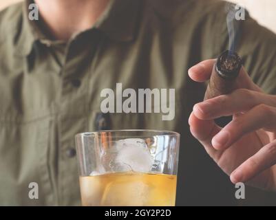 Ein Mann in einem grünen Hemd hält eine kubanische Zigarre und ein Glas Whiskey auf Eis. Nahaufnahme einer rauchenden Zigarre. Stockfoto