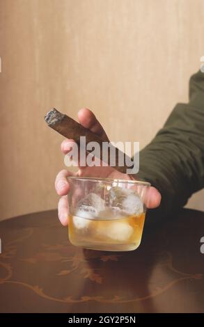 Ein Mann in einem grünen Hemd hält eine kubanische Zigarre und ein Glas Whiskey auf Eis. Nahaufnahme einer rauchenden Zigarre. Stockfoto