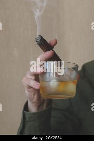 Ein Mann in einem grünen Hemd hält eine kubanische Zigarre und ein Glas Whiskey auf Eis. Nahaufnahme einer rauchenden Zigarre. Stockfoto