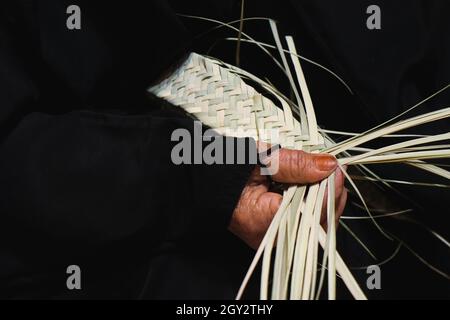 Hände älterer arabischer Frau, die in traditionellem schwarzen Kleid in Nahaufnahme einen Korb aus getrockneten Bio-Palmblättern webt. Stockfoto