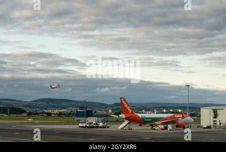 Ein easyJet Airbus A320-200 mit einer Europcar-Anzeige parkte auf dem Vorfeld am Flughafen Glasgow, Schottland, während ein weiteres Easyjet-Flugzeug abhebt. Stockfoto