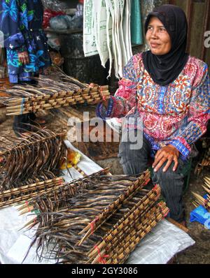 Eine Frau auf einem Markt im Freien, die in einer bunten Bluse sitzt und einen schwarzen Hijab trägt, der geräucherte Aale in Bukittinggi, West Sumatra, Indonesien verkauft. Stockfoto