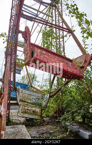 Verlassene, rot-weiße und blaue Sitze mit grünen Büschen auf einem Riesenrad in einem geschlossenen Freizeitpark Stockfoto