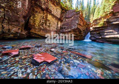 Rote und blaue Flussfelsen im flachen Fluss am Fuße der Schlucht mit Wasserfall und steilen Klippen Stockfoto