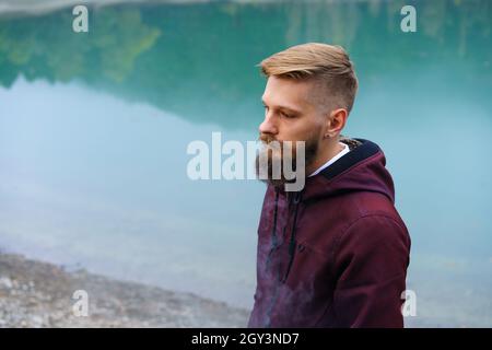Der junge bärtige Mann stand allein am See und schaute nachdenklich rauchend zur Seite. Nebel über dem Wasser. Neblige Luft. Früher kühler Morgen. Ein dunkler, beängstigender Moment und eine dunkle Atmosphäre. Stockfoto