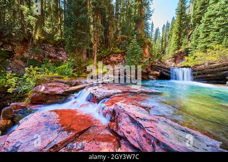 Majestätische Wasserfälle über roten Felsen in einer Schlucht aus moosigen Felsen und Kiefern Stockfoto