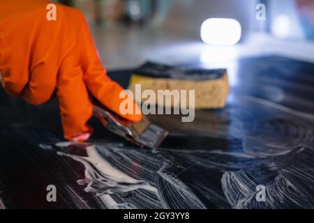 Nahaufnahme eines orangefarbenen Handschuhs, eines Metallschabers und eines Schwamms, der das Kochfeld mit einem Reinigungsmittel wäscht. Selektiver Fokus. Das Konzept der Hausreinigung eines elektrischen Stockfoto