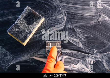 Draufsicht. Ein orangefarbener Handschuh, ein Metallschaber und ein Schwamm reinigen das Kochfeld mit Reinigungsmittel. Das Konzept der Hausreinigung ein elektrischer Stow Stockfoto
