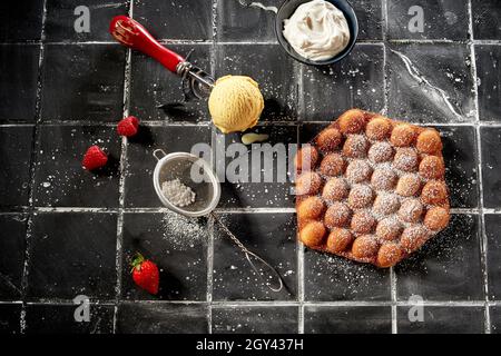 Ein leckeres Sommerdessert mit Blasenwaffeln und Eis in der Ansicht von oben auf der goldenen Eierwaffel, einer Kugel, einem Sieb mit Zucker und einer Schüssel mit Peitsche zubereiten Stockfoto